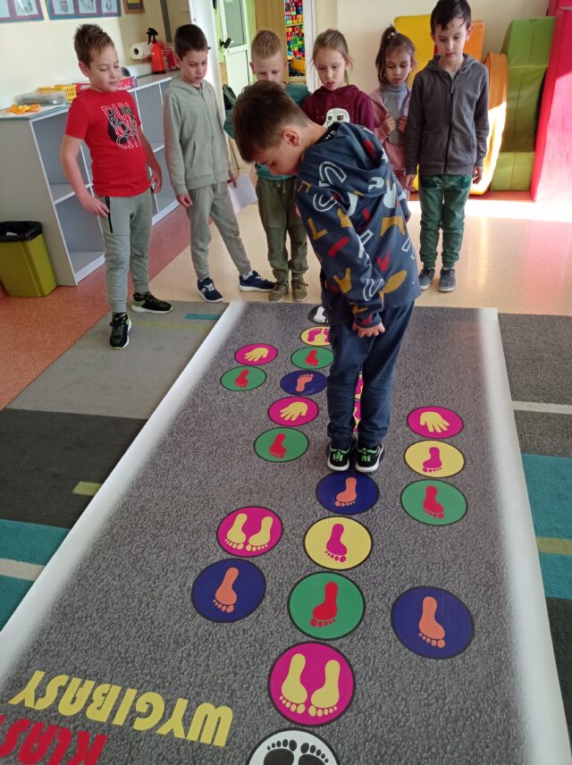 Siedmioro dzieci stoi przy macie podłogowej do gry „Klasy – wygibasy”. Igor przeskakuje na kolejne pole w grze.