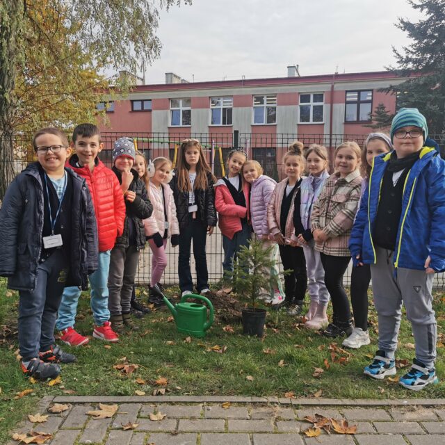 Grupa uczniów stoi na tle drzewa liściastego.